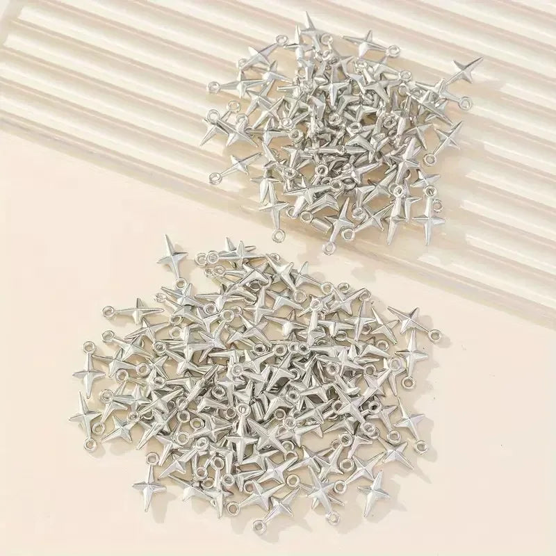 A pile of small silver metal four-pointed star charms with loops at the ends, arranged on a light beige surface with a cream ribbed background.