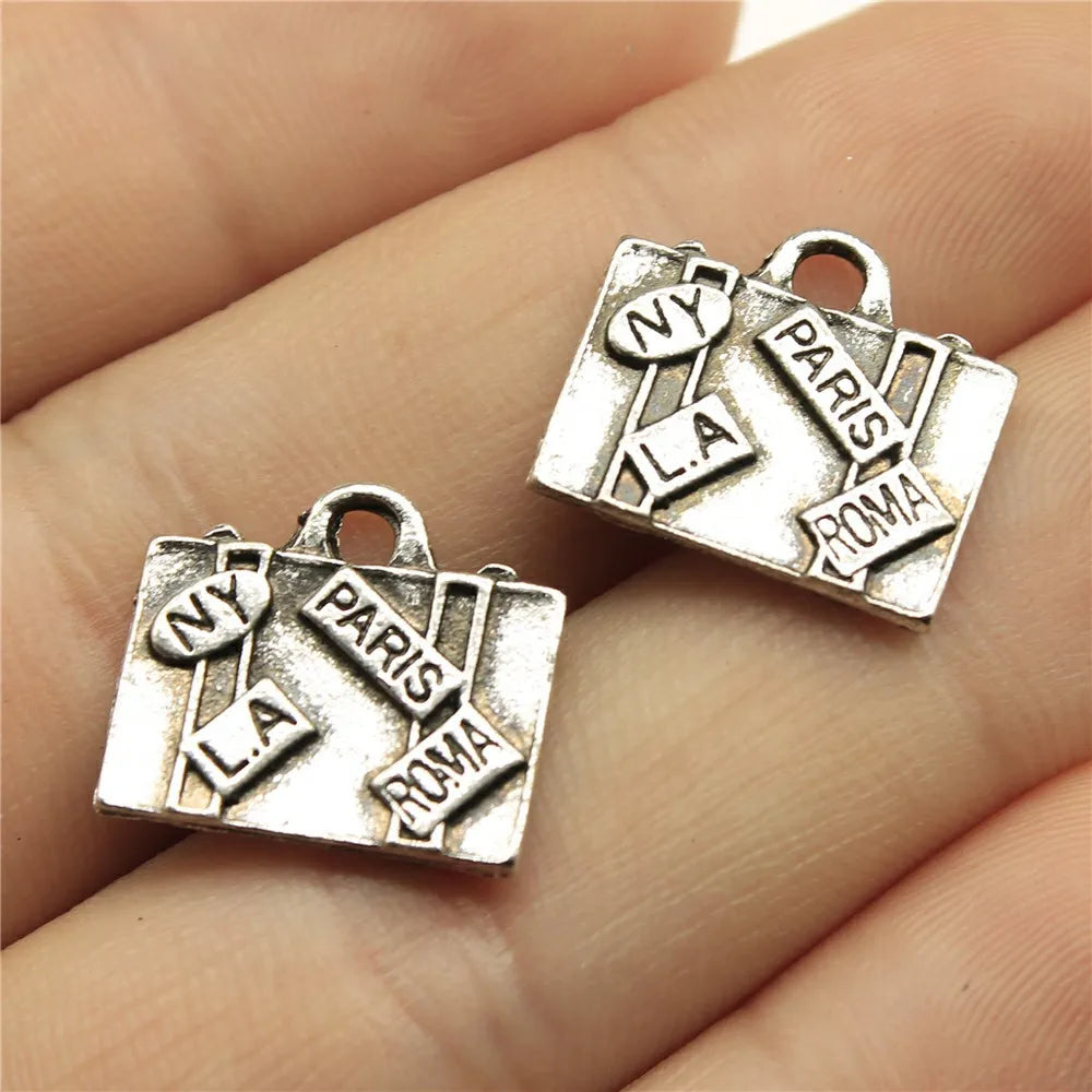 Two silver-toned jewelry charms shaped like vintage suitcases with travel city names (NY, LA, PARIS, ROMA) engraved on them, displayed on a person's fingers.