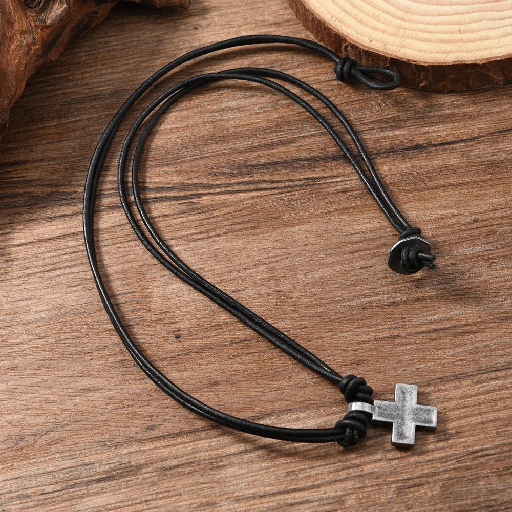 A black cord necklace with a silver-toned cross pendant, displayed on a rustic wooden surface.