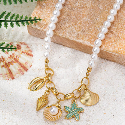 Pearl necklace with gold and turquoise shell charms on a sandy surface with shells and a rock.