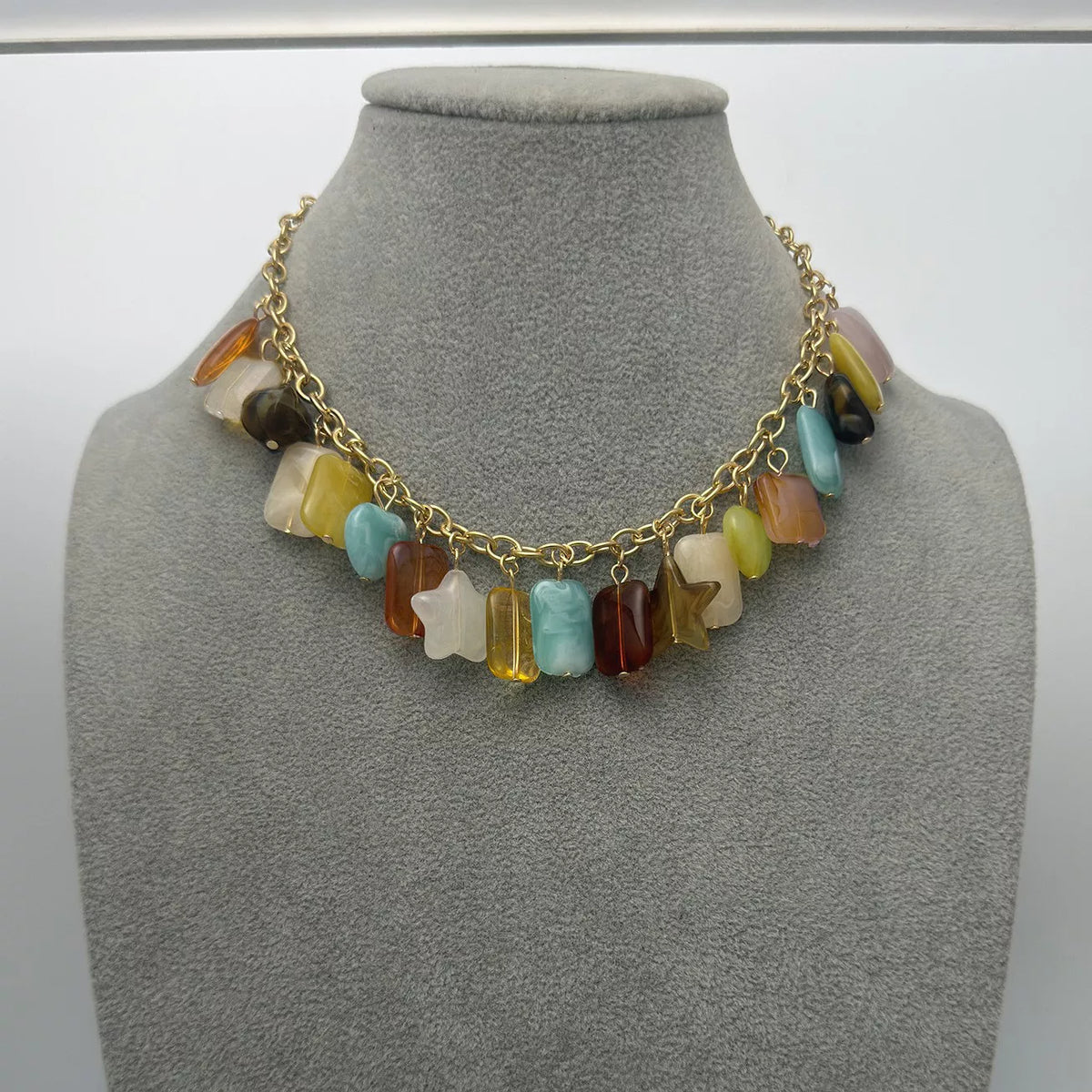 A gold chain necklace with a row of colorful, irregularly shaped stone beads and two star-shaped charms, displayed on a gray fabric jewelry bust.