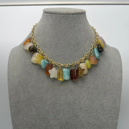 A gold chain necklace with a row of colorful, irregularly shaped stone beads and two star-shaped charms, displayed on a gray fabric jewelry bust.