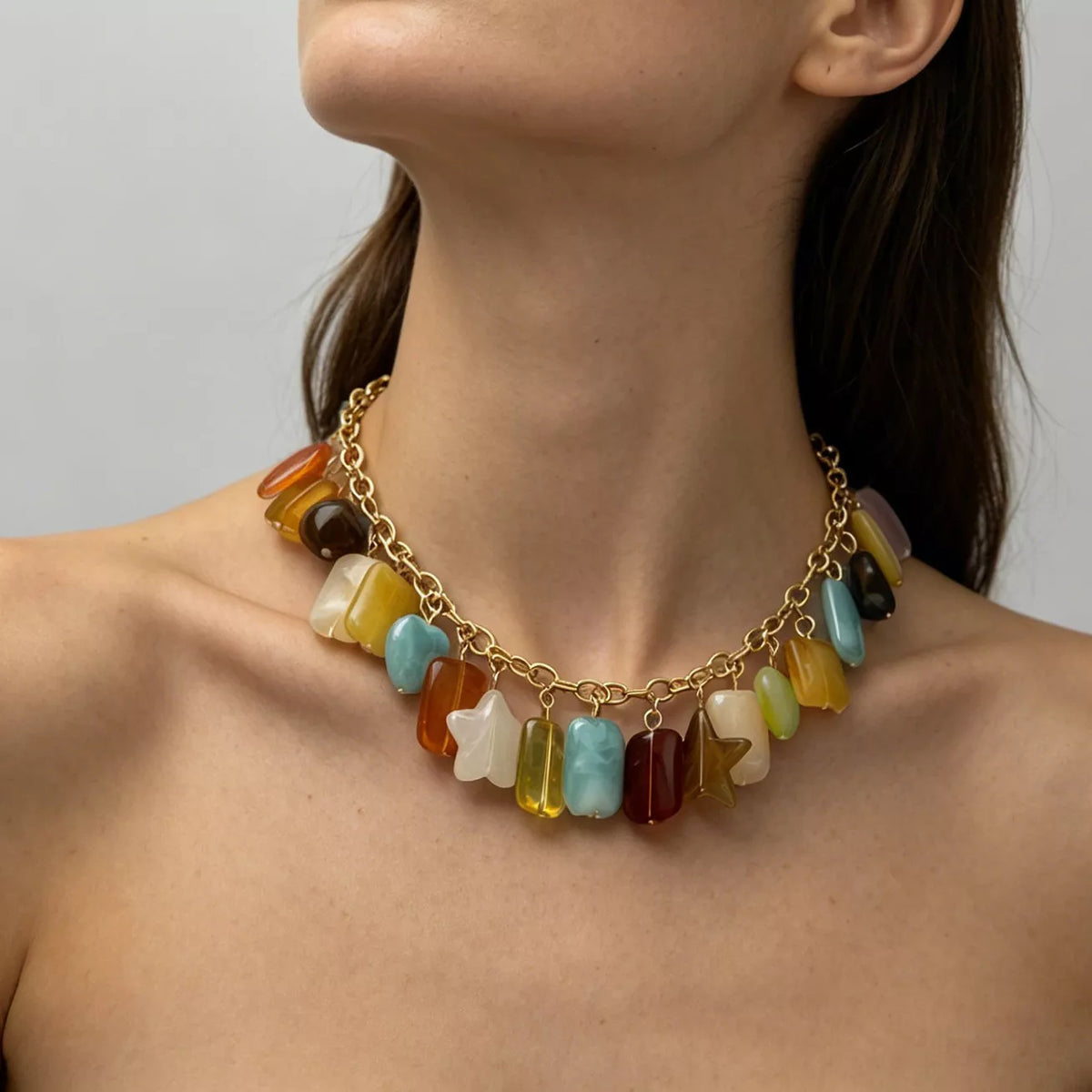 A close-up of a model wearing a gold chain necklace adorned with colorful, irregularly shaped stone-like charms in yellow, blue, orange, brown, and white, set against a neutral gray background.