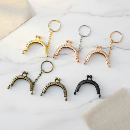 Six antique-style kiss-lock coin purse keychains in different metal finishes (gold, silver, rose gold, bronze, and black) arranged on a plain white background.