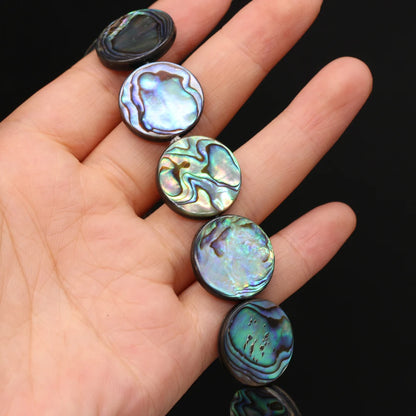 A hand holding a strand of round abalone shell beads with iridescent green, blue, and purple patterns against a black background.
