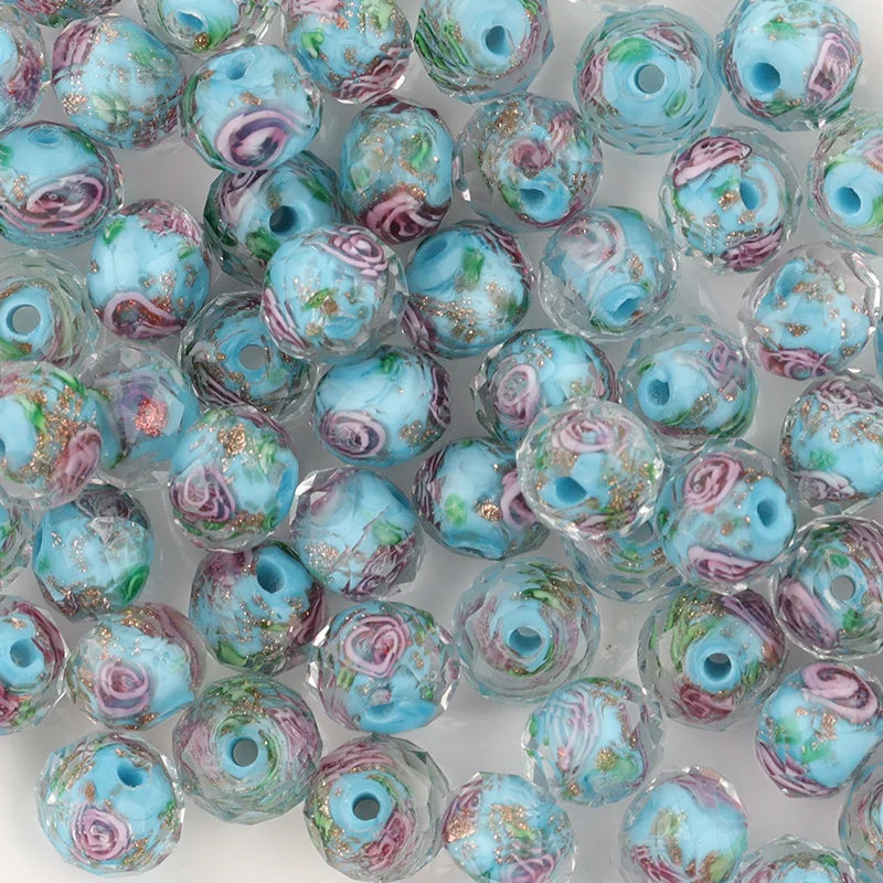 A pile of round glass beads with a light blue and white marbled base, decorated with pink rose motifs, green leaves, and gold foil flecks.