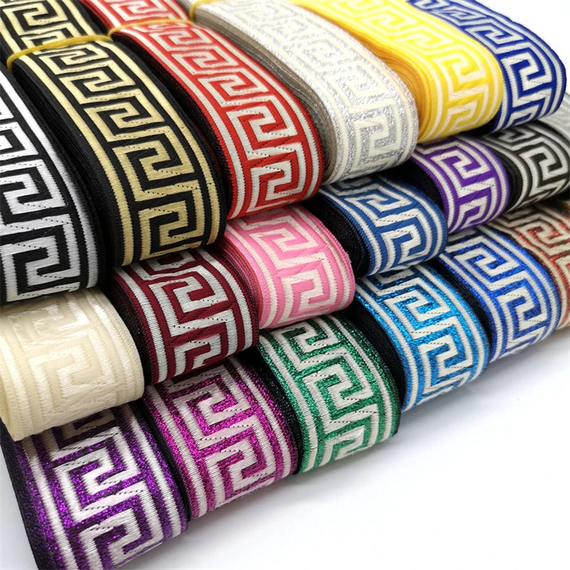 A close-up of multiple rolls of decorative trim with a Greek or vintage boho key pattern, shown in black, gold, red, white, yellow, blue, pink, purple, green, and glittery purple tones on a white background.