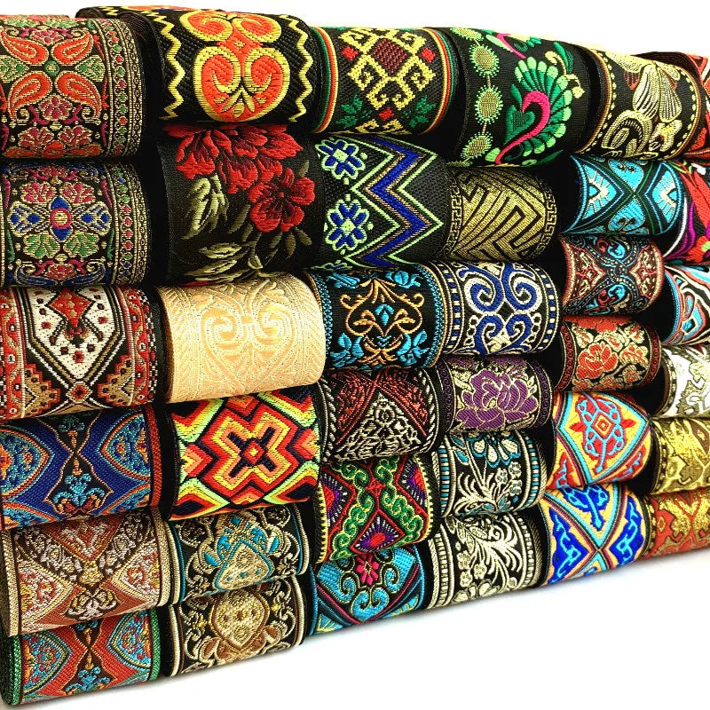 A stack of colorful embroidered ribbon trim rolls, bohemian style, with intricate geometric and floral patterns in red, blue, green, yellow, black, and gold, shown against a white background.