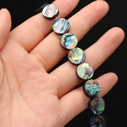 A close-up of a hand holding a strand of round abalone shell beads with an iridescent, multicolored pattern against a black background.