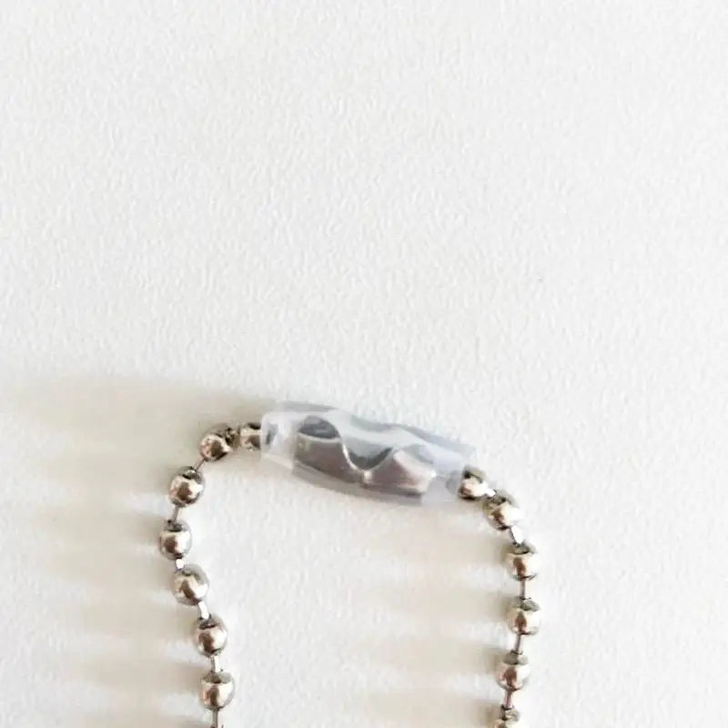 A close-up of a silver beaded chain with a translucent smoky gray oval charm, resting on a plain white textured surface.