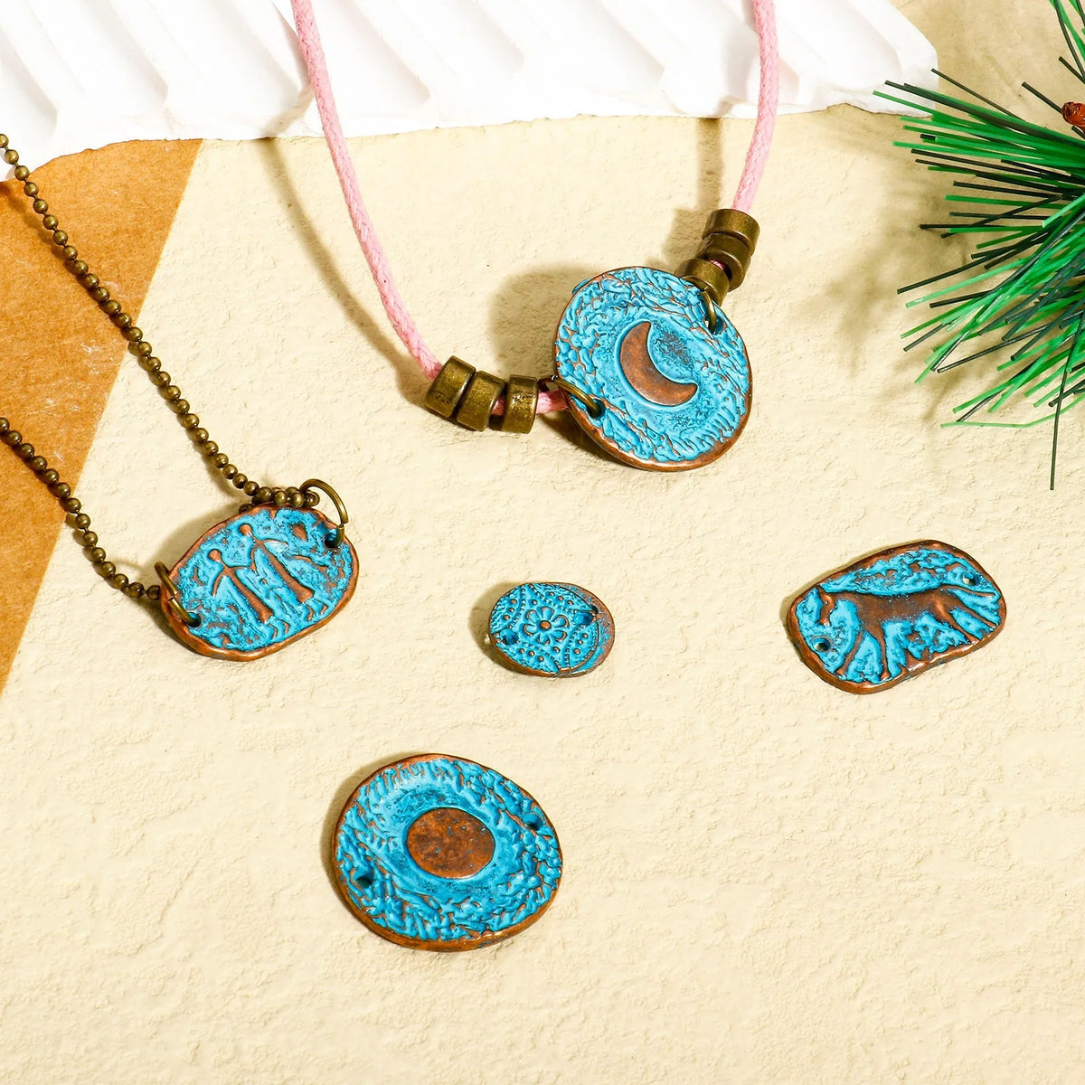 A set of turquoise and bronze pendant necklaces and charms displayed on a textured beige surface with a pink cord necklace and small decorative pine branches.