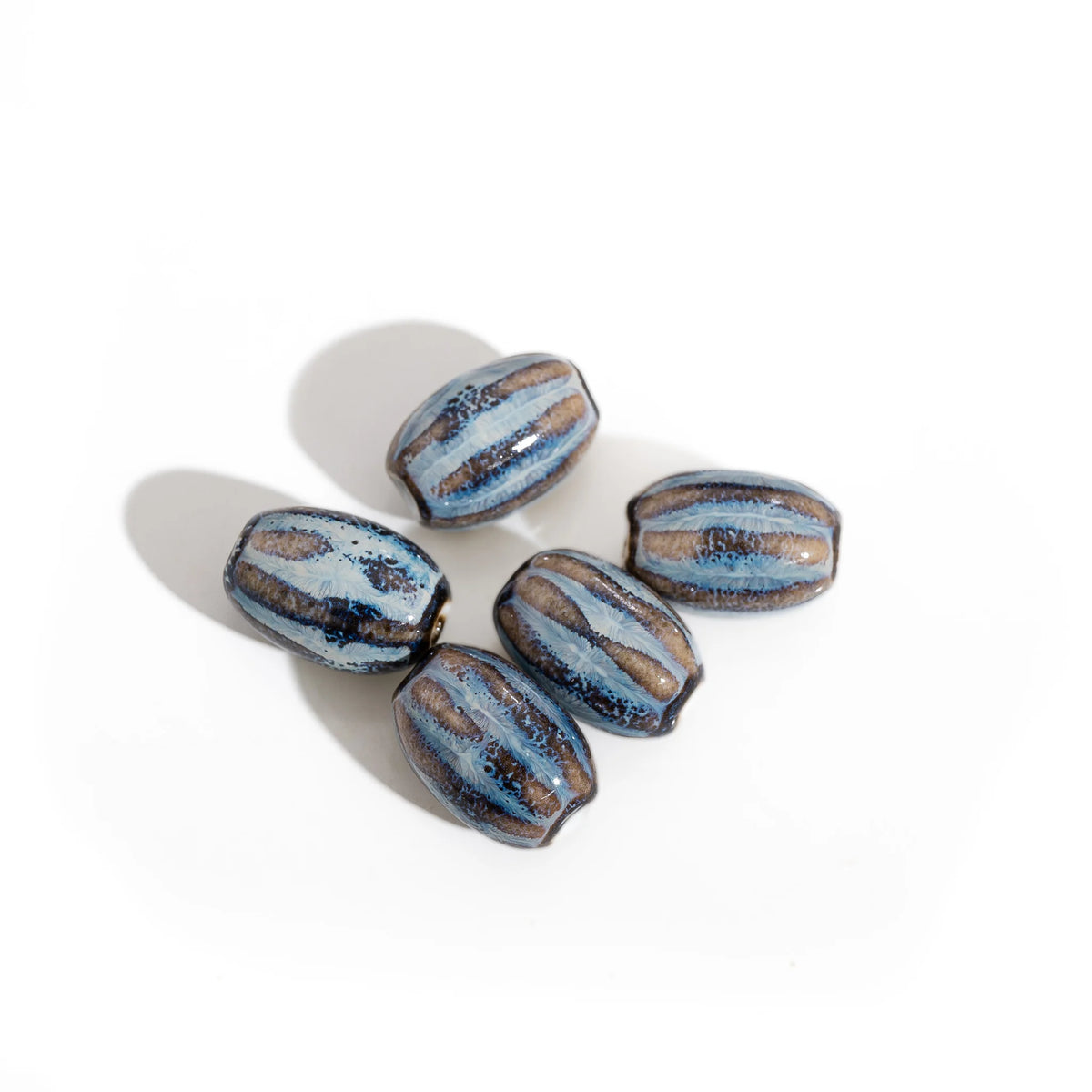 Five handmade ceramic beads with a glossy finish, featuring a blue and brown striped pattern with dark metallic accents, arranged on a white background with soft shadows.