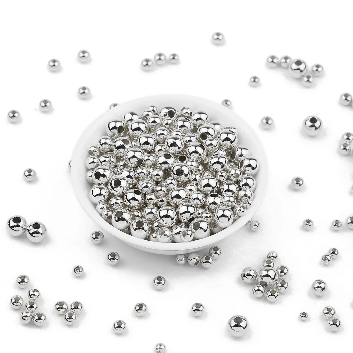 Silver metallic round beads scattered around a small white bowl on a white background.