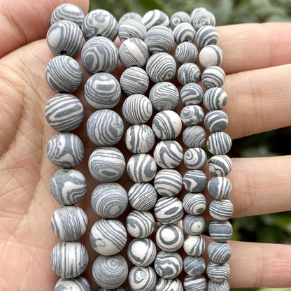 Multiple strands of round beads with a gray and white swirling stone pattern, held in a person's hand against a blurred green background.