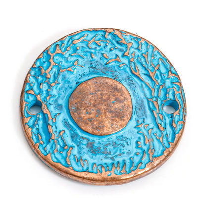 A round ceramic pendant with a textured turquoise blue glaze and a central copper-toned medallion, featuring two side holes, shown on a white background.