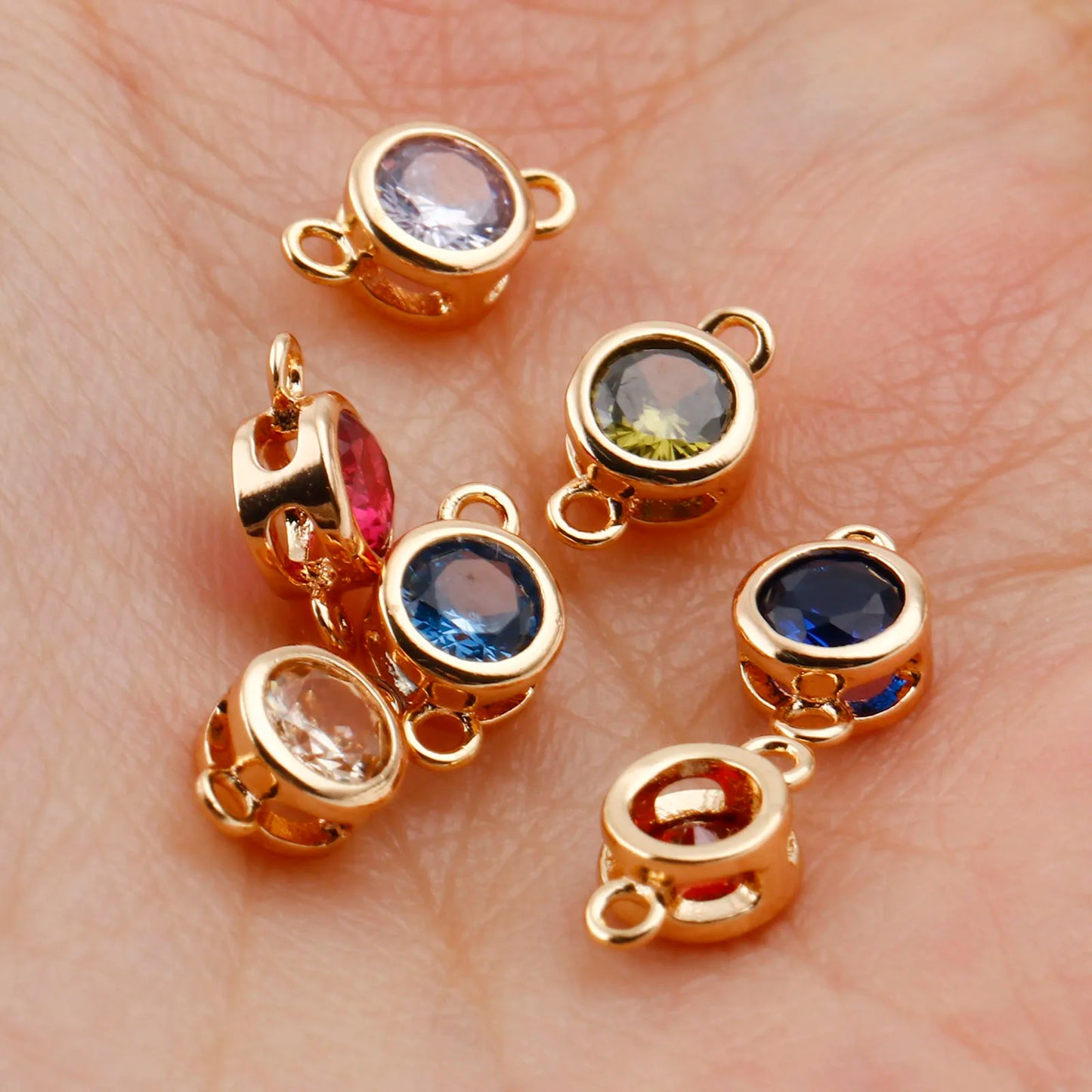 Six gold-plated round bezel charm links with various colored gemstones, resting on a person's palm to show scale.