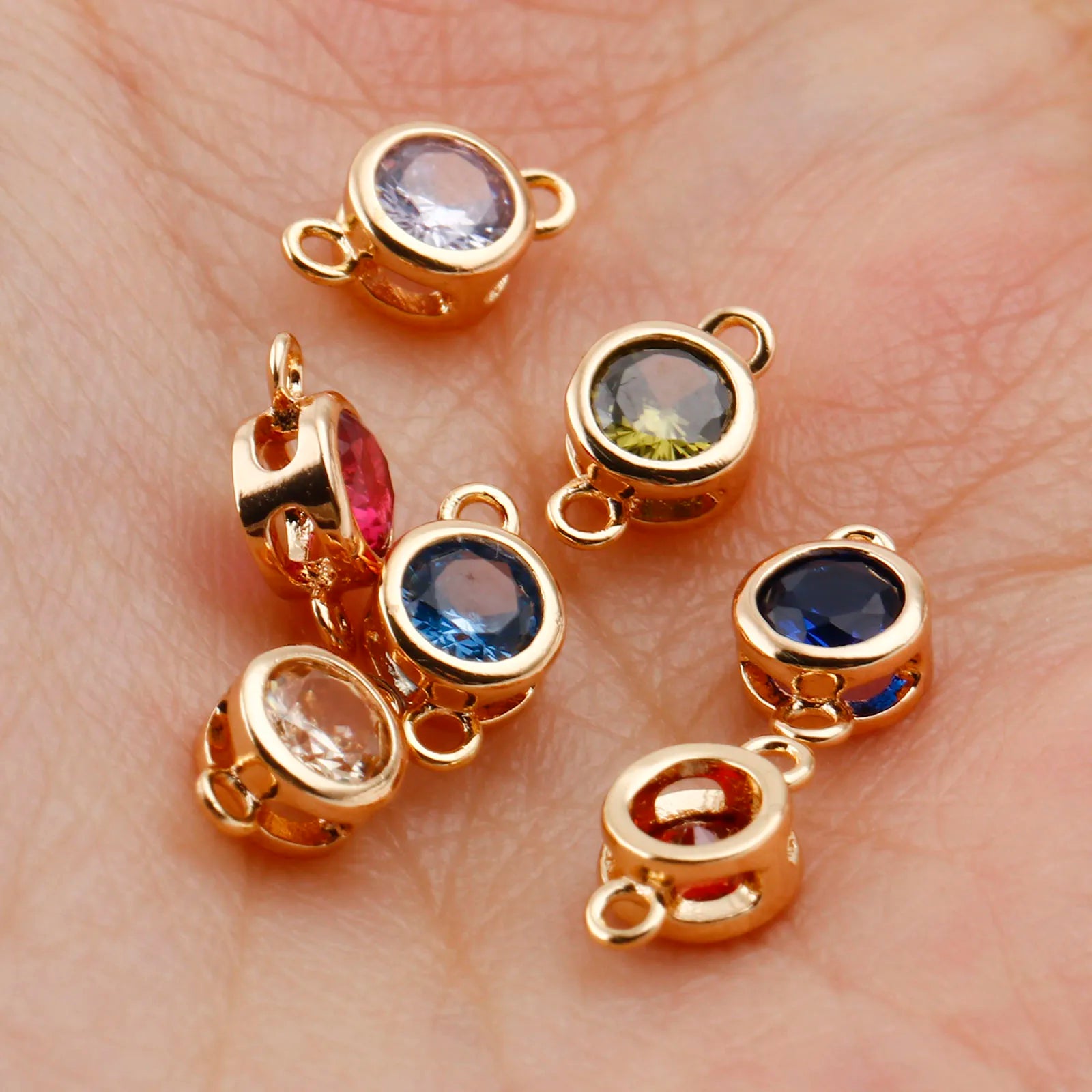 Six gold-plated round bezel charm links with various colored gemstones, resting on a person's palm to show scale.