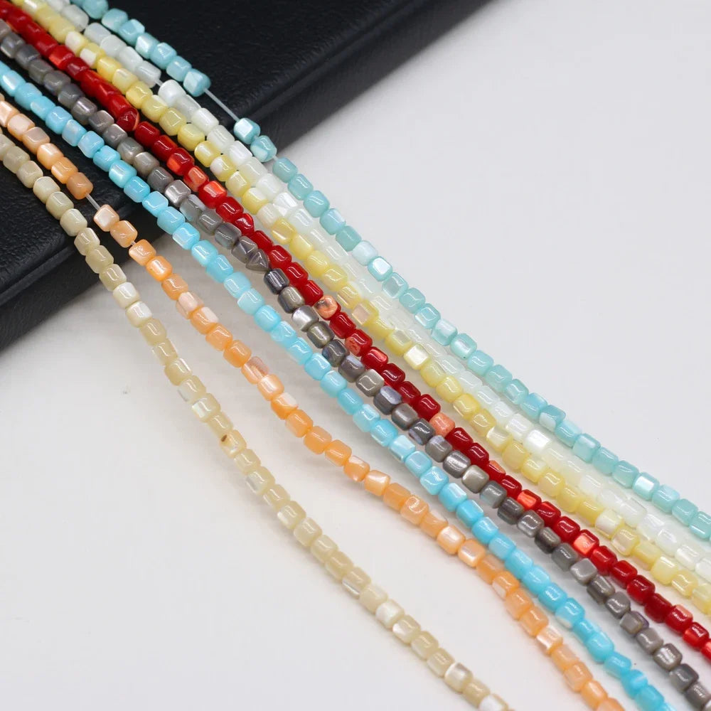 Multiple strands of small faceted shell beads in assorted colors, including light blue, red, gray, yellow, peach, and cream, arranged diagonally on a white surface with a black textured block in the background.