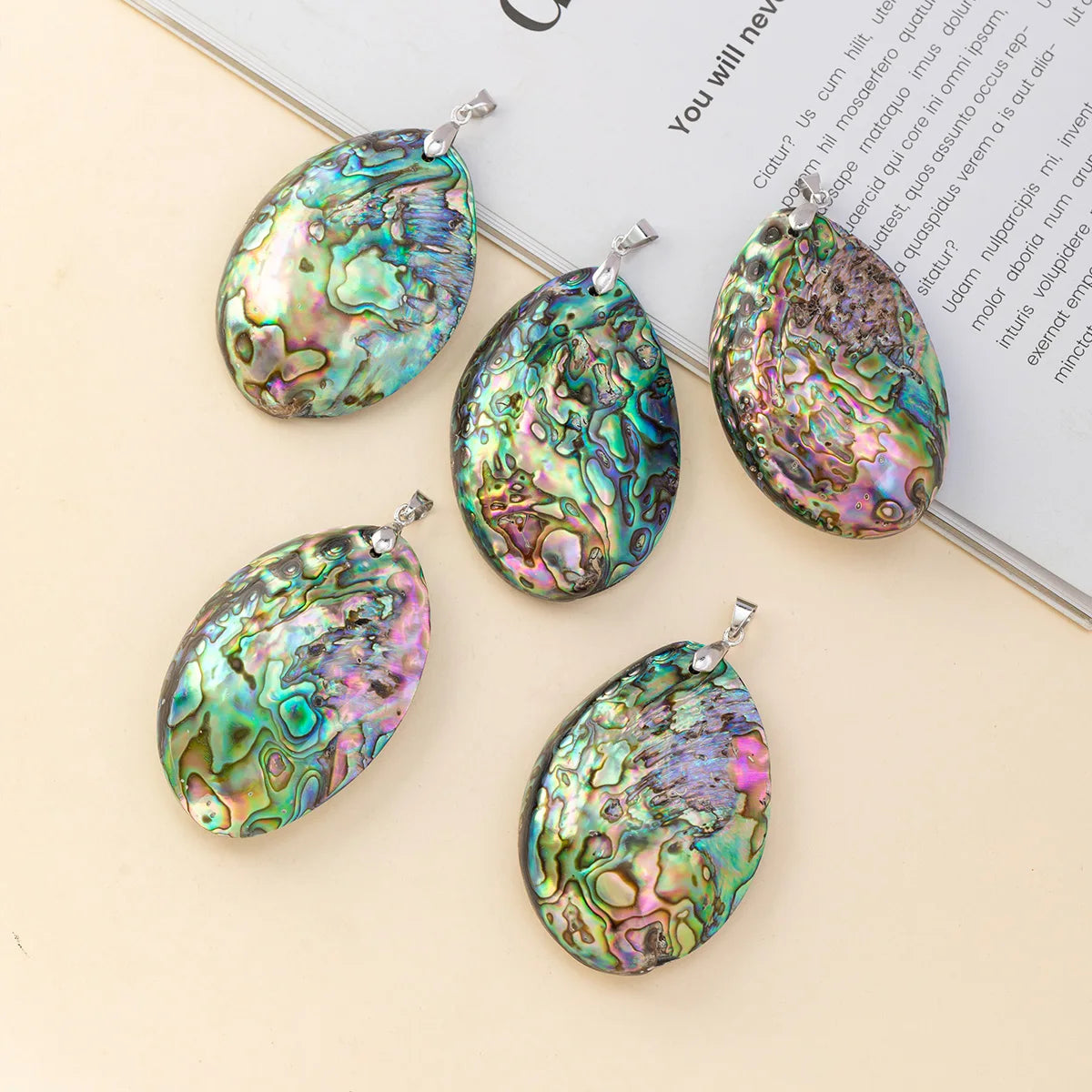 Five teardrop-shaped abalone shell pendants with silver bail loops, displayed on a light beige surface next to an open book.