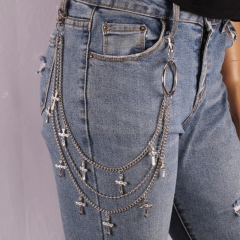 Silver-tone metal chain accessories with multiple cross pendants and a large ring, draped over light-wash blue jeans on a model.