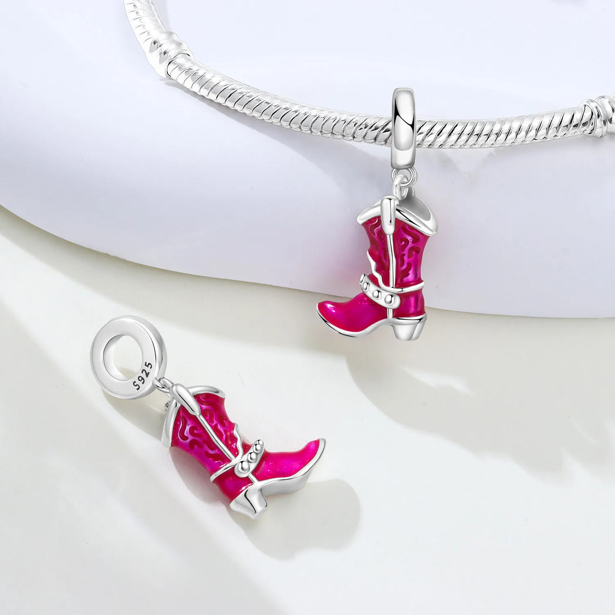 A pair of silver cowboy boot charms with bright pink enamel detailing, shown with one charm on a white display stand and the other floating below. A silver snake chain bracelet is draped across the top of the stand.
