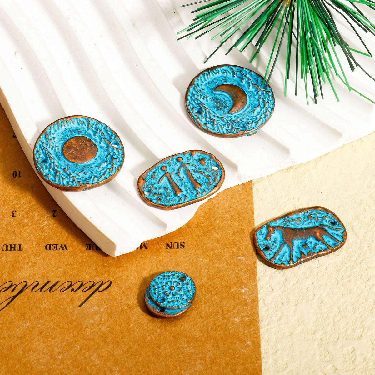 Five turquoise-blue carved wooden beads with rustic brown edges, featuring various symbols and designs, arranged on a styled flat-lay background of white slatted paper, kraft paper, and a green pine branch.