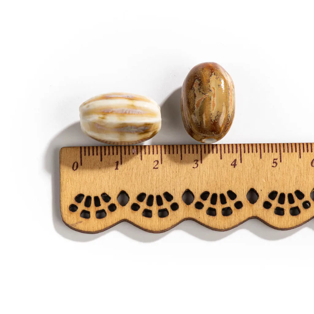 Two small oval beads, one striped cream and brown and one polished brown with natural marbling, shown next to a wooden ruler with a scalloped edge on a white background.
