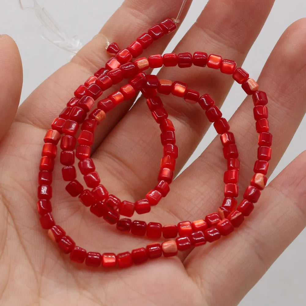 A hand holding a strand of small red faceted shell beads, with some translucent pale pinkish-red beads mixed in, against a plain light background.