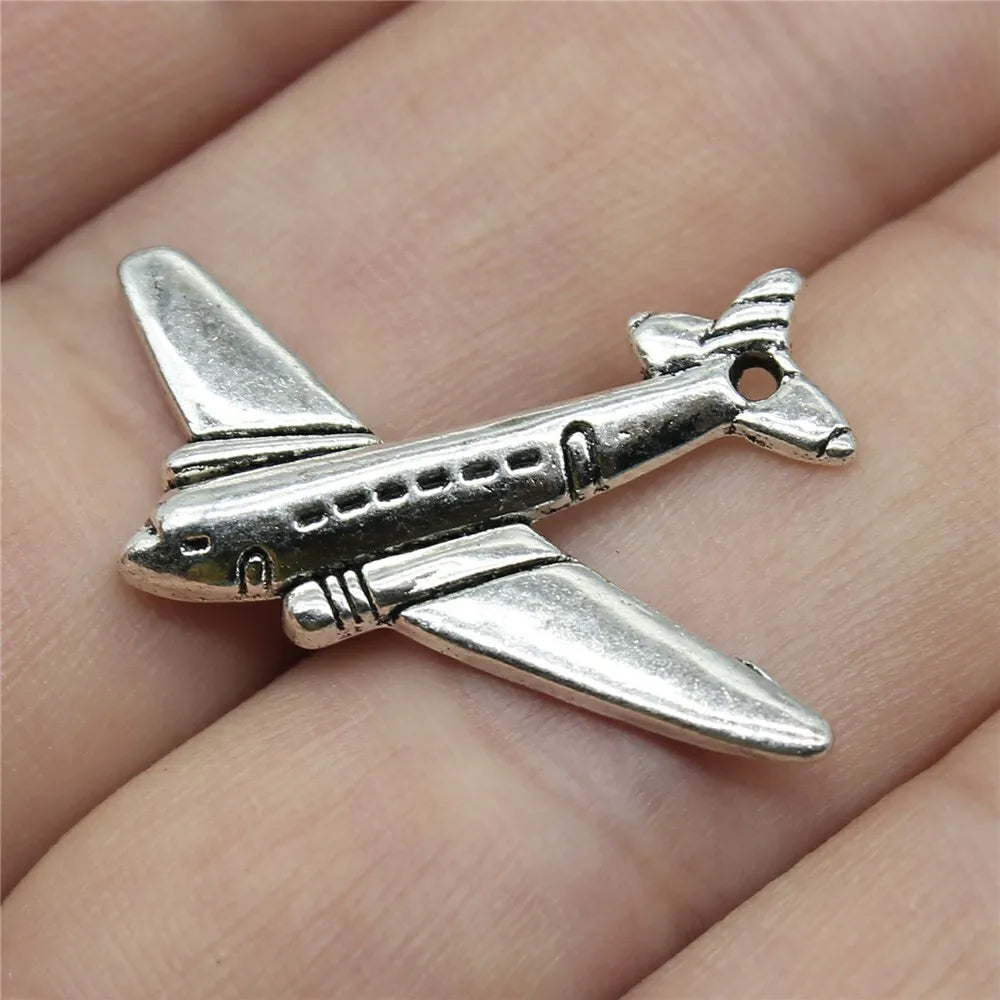 Silver airplane pendant on a person's finger. Travel charms for jewelry making featuring airplane, passport, suitcase and camera charms.