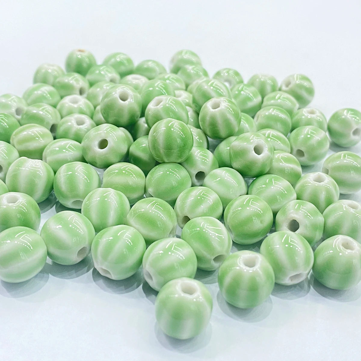 A pile of glossy, light green ceramic beads with a ribbed, pumpkin-like texture and a central hole, arranged on a clean white background.