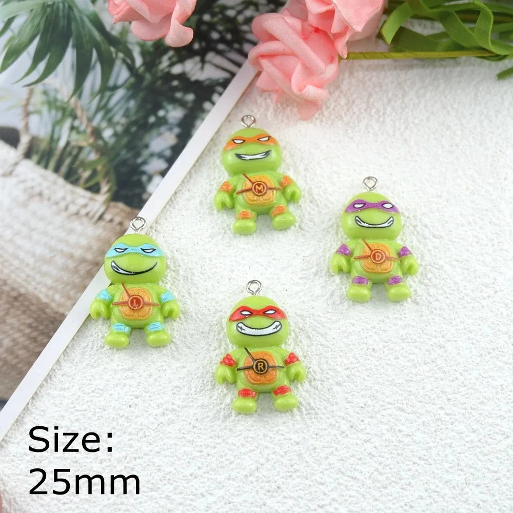Four small green cartoon turtle charms with colorful masks and metal loops, arranged on a white textured surface with decorative flowers and a plant in the background. Text in the lower left reads 'Size: 25mm'.