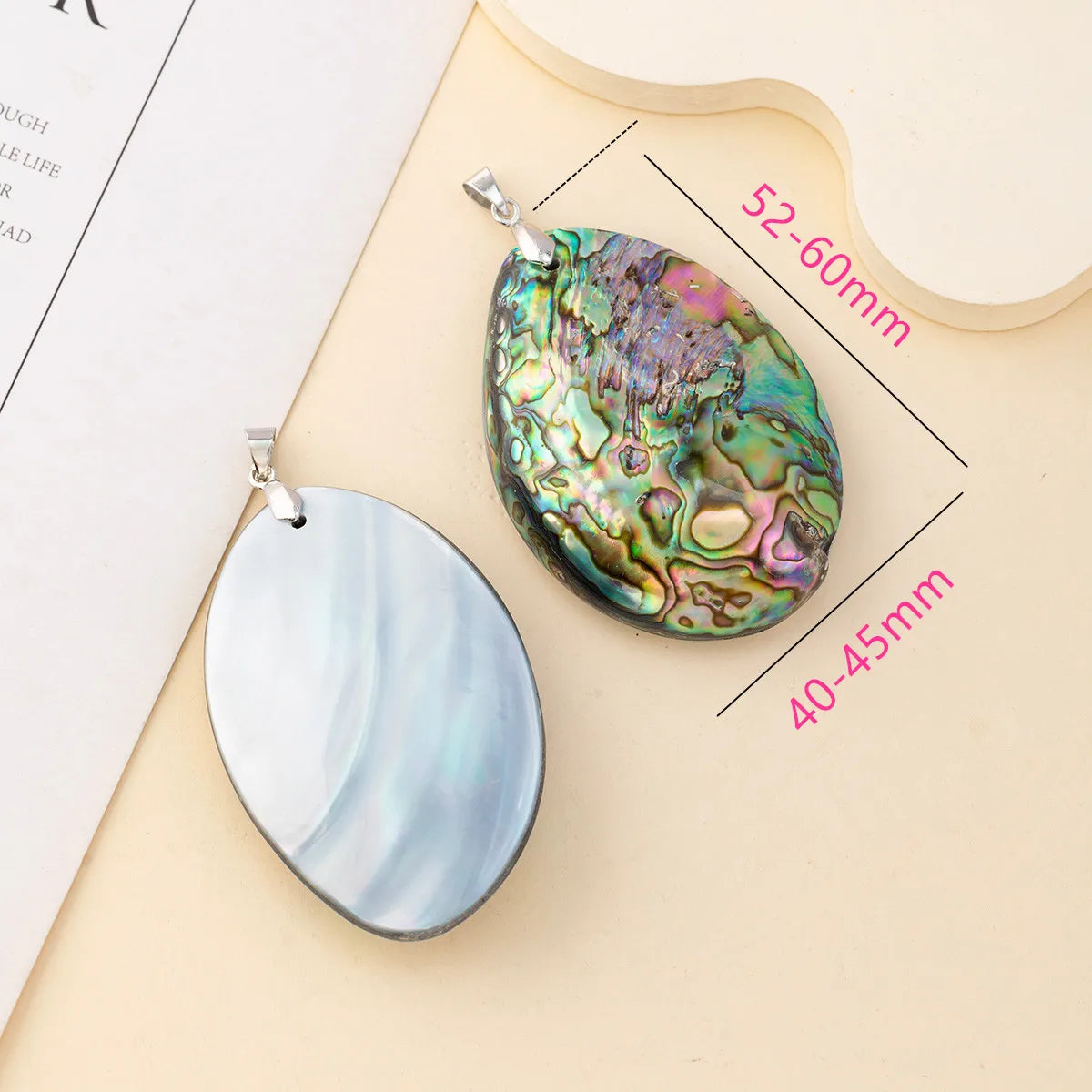 Two teardrop-shaped abalone shell pendants displayed on a beige surface, one with a silver bail and a multicolored iridescent shell, and one with a silver bail and a pale blue-gray shell. Pink text indicates dimensions: 52-60mm and 40-45mm.