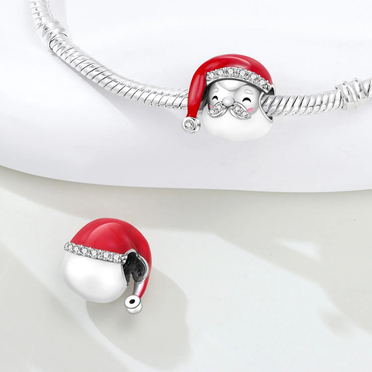A silver-toned snake chain bracelet with a Santa hat charm, shown on a white surface with a matching charm detached below. The Santa hat charm has red and white enamel, a silver beaded brim, and a small pink accent near the eye.
