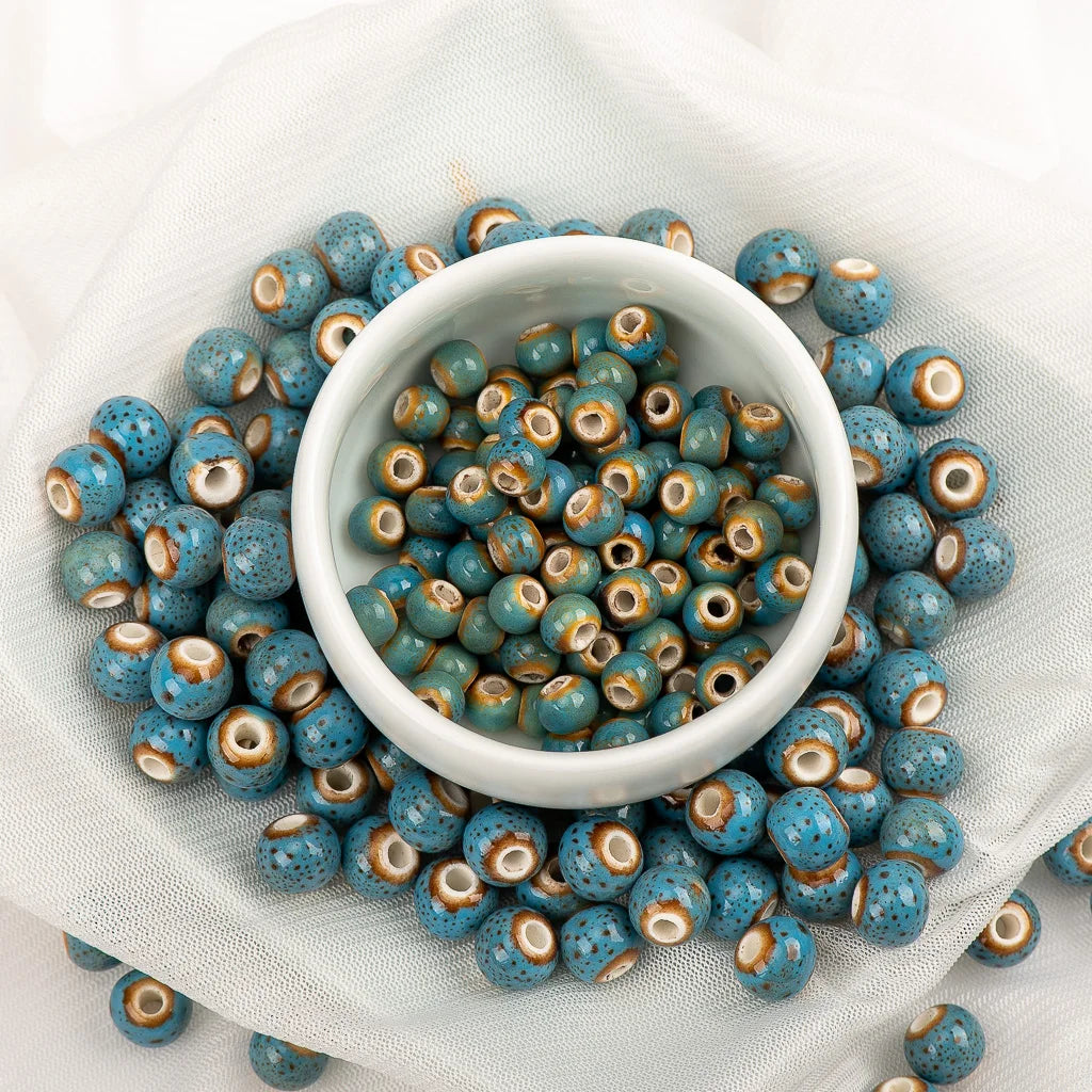 Blue speckled seed beads with brown and cream rings, arranged around a small white bowl on a white fabric background.