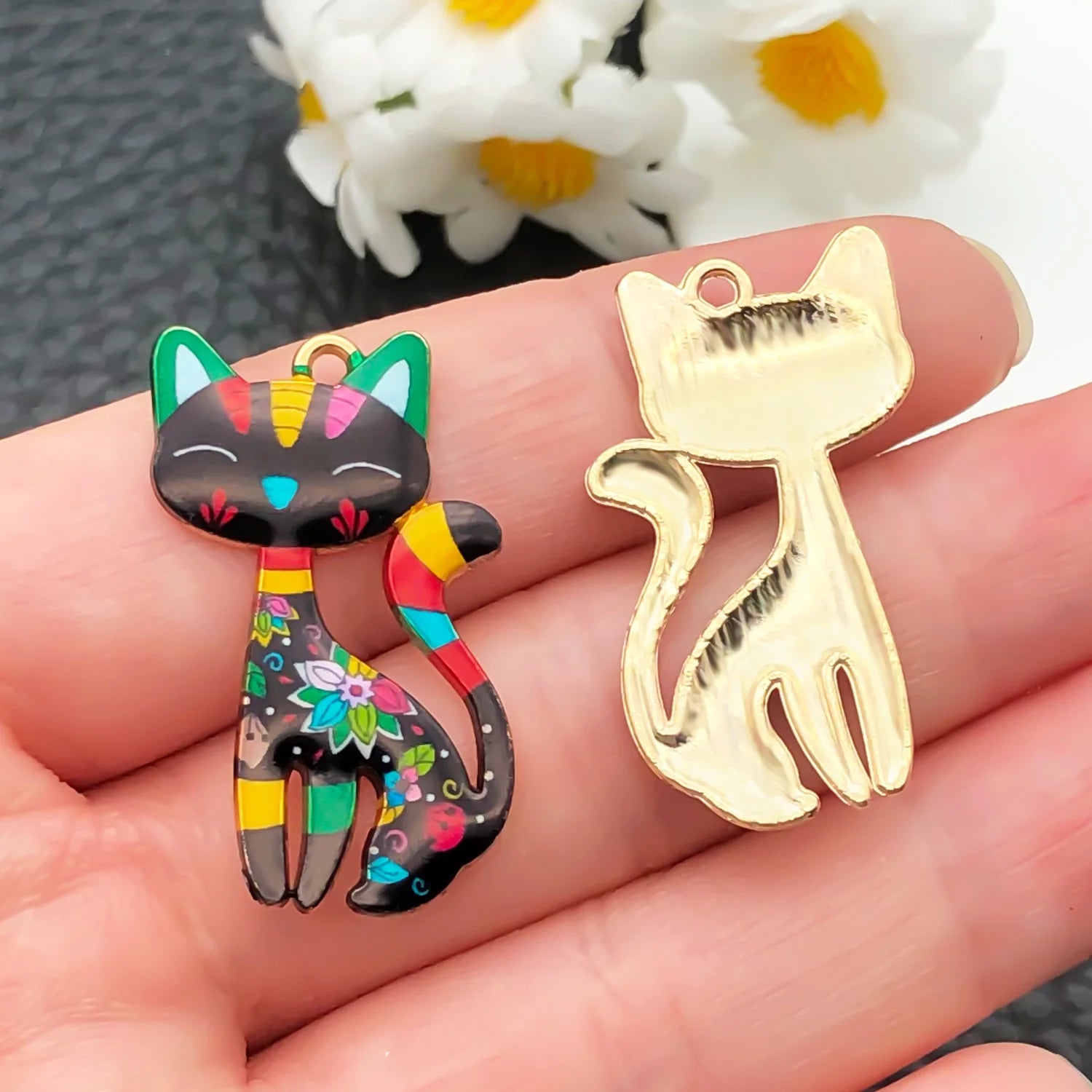 Colorful cat-shaped charm and gold cat-shaped pendant on a hand with flowers in the background.