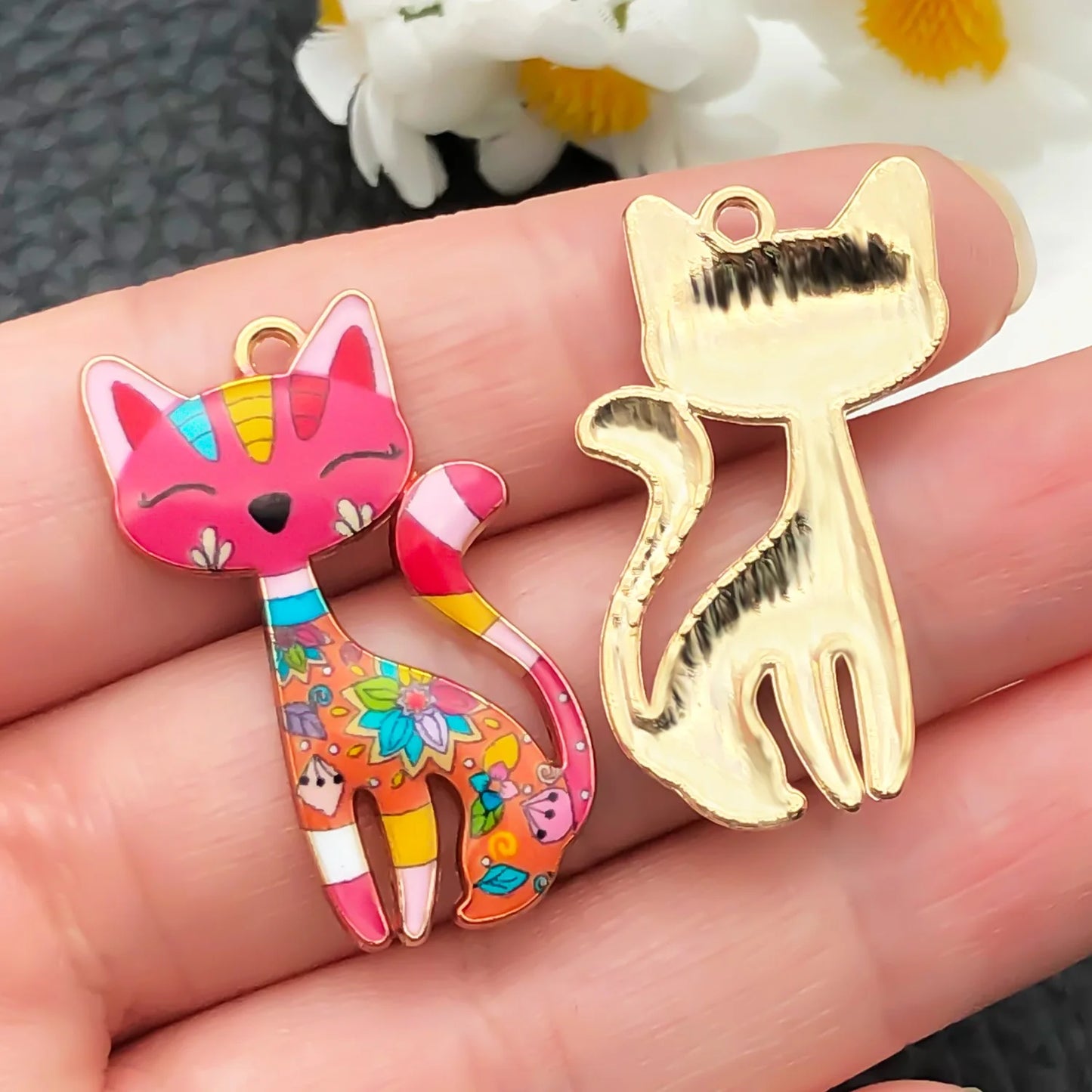 Colorful cat-shaped charm and gold cat-shaped charm held in a hand with flowers in the background.