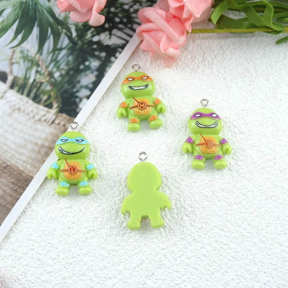 Four small green cartoon turtle charms with colorful masks and silver loops, plus one plain green silhouette charm, arranged on a white textured surface with pink flowers in the background.