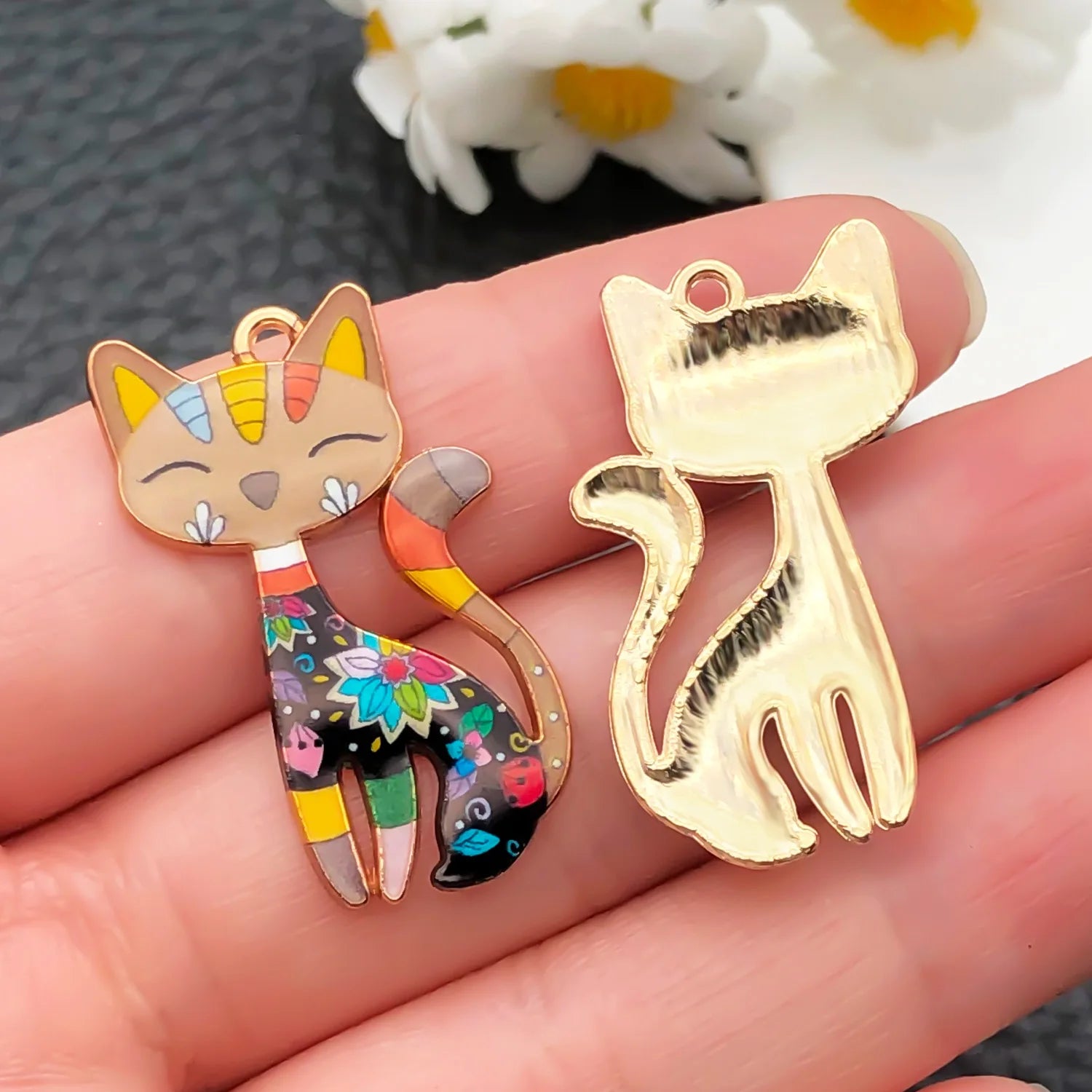 Two cat-shaped pendants, one colorful and one gold, held between fingers with a blurred floral background.