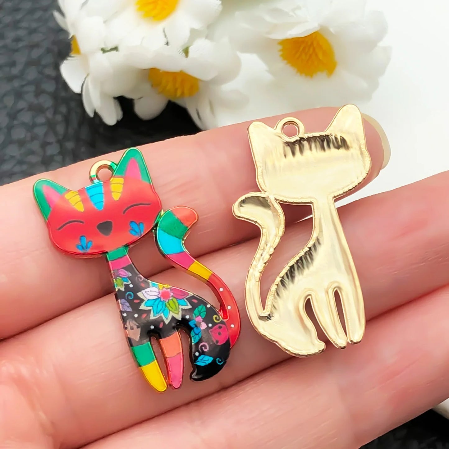 Colorful cat-shaped charm and gold cat-shaped pendant on a hand with flowers in the background.