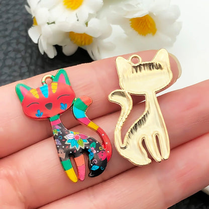 Colorful cat-shaped charm and gold cat-shaped pendant on a hand with flowers in the background.