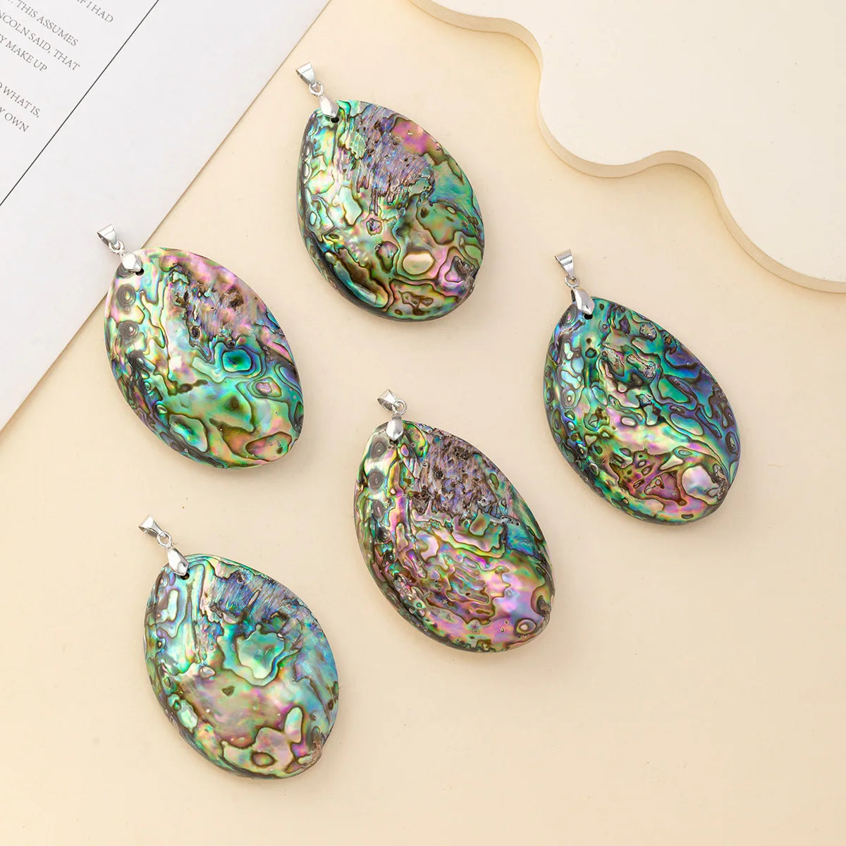 Five oval abalone shell pendants with silver bail loops, arranged on a beige background with a decorative paper and wooden slice prop.