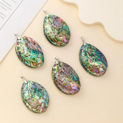 Five oval abalone shell pendants with silver bail loops, arranged on a beige background with a decorative paper and wooden slice prop.