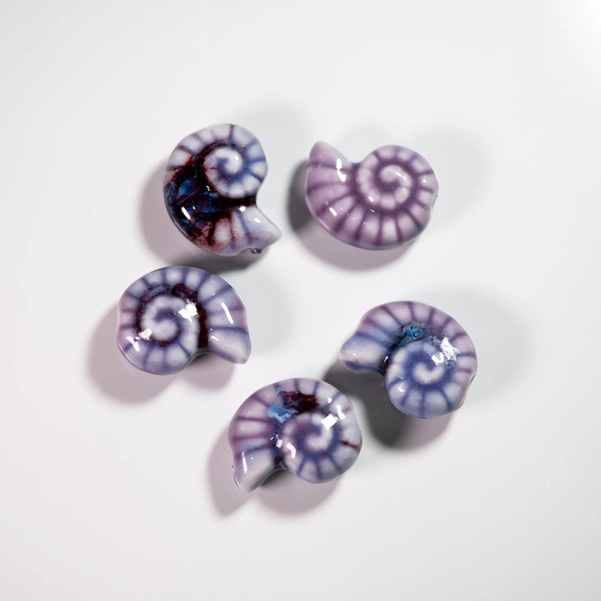 Five glossy spiral conch shell-shaped beads in shades of purple, lavender, and white, with one featuring dark blue and red marbling, arranged on a plain white background.