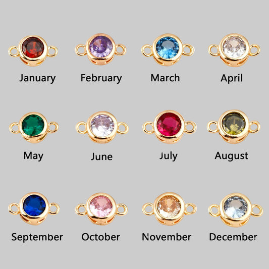 Twelve gold-toned round bezel settings with small side loops, each labeled January through December, shown on a white background. The stones change color monthly: red, purple, blue, clear/white, green, pale lavender, pink, olive green, dark blue, pale pink, peach, and light blue.