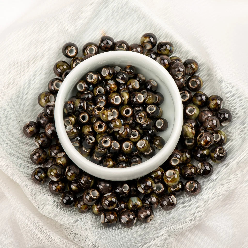 Dark brown and black glossy glass beads with white center holes, arranged around a small white ceramic bowl on a white fabric background.
