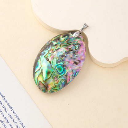 An oval abalone shell pendant with a silver bail, displayed on a light beige background with a decorative wooden slice and a corner of a book visible.