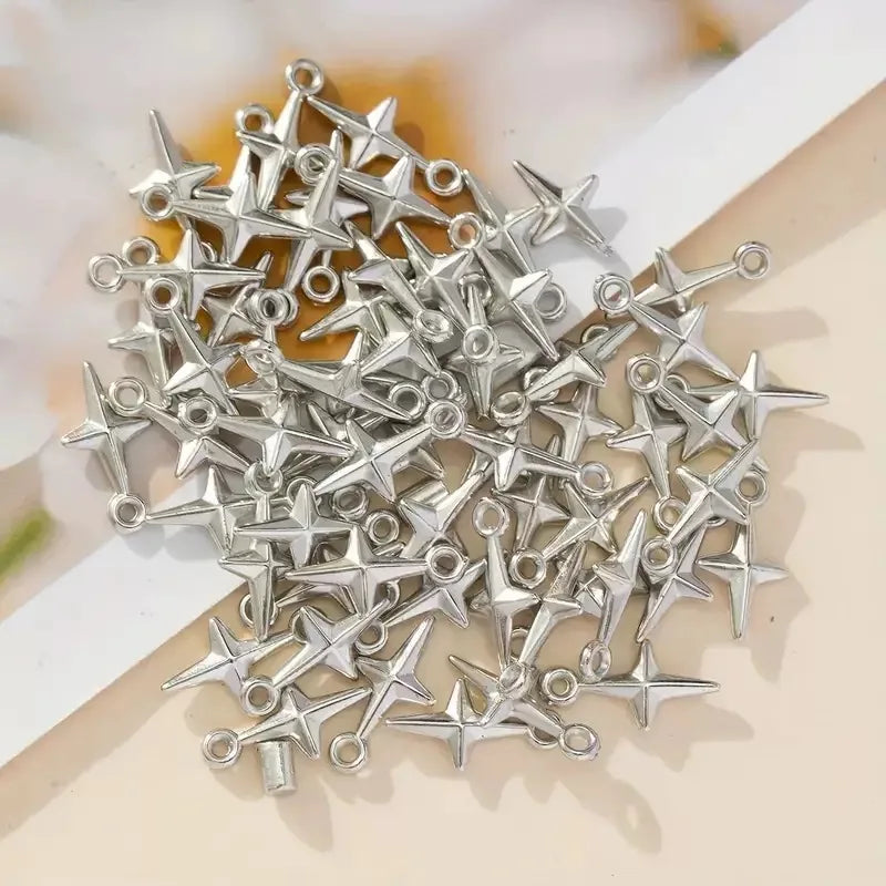 A pile of small silver-toned metal star-shaped charms with loop tops, photographed on a light beige surface with a white ribbon.