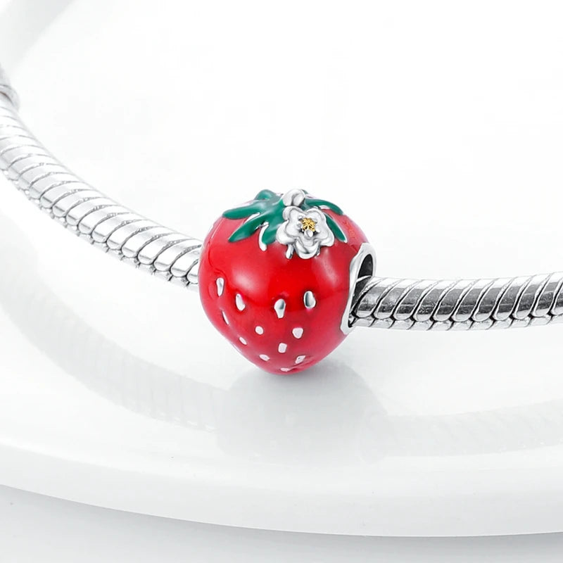 A close-up of a red strawberry charm with green leaves and a white flower detail, threaded onto a silver snake chain bracelet against a clean white background.