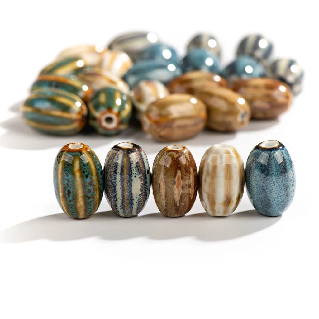 Five glossy ceramic bead spacers with vertical striped patterns in earthy tones of blue, brown, beige, and green, arranged in a row with a larger, out-of-focus group in the background on a white surface.