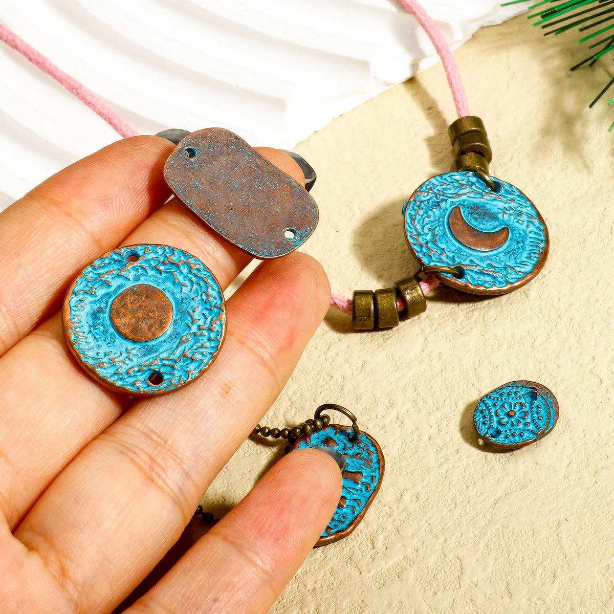 A hand holds a round turquoise-blue and bronze pendant necklace charm, with additional matching charms and a pink cord laid out on a textured beige surface.